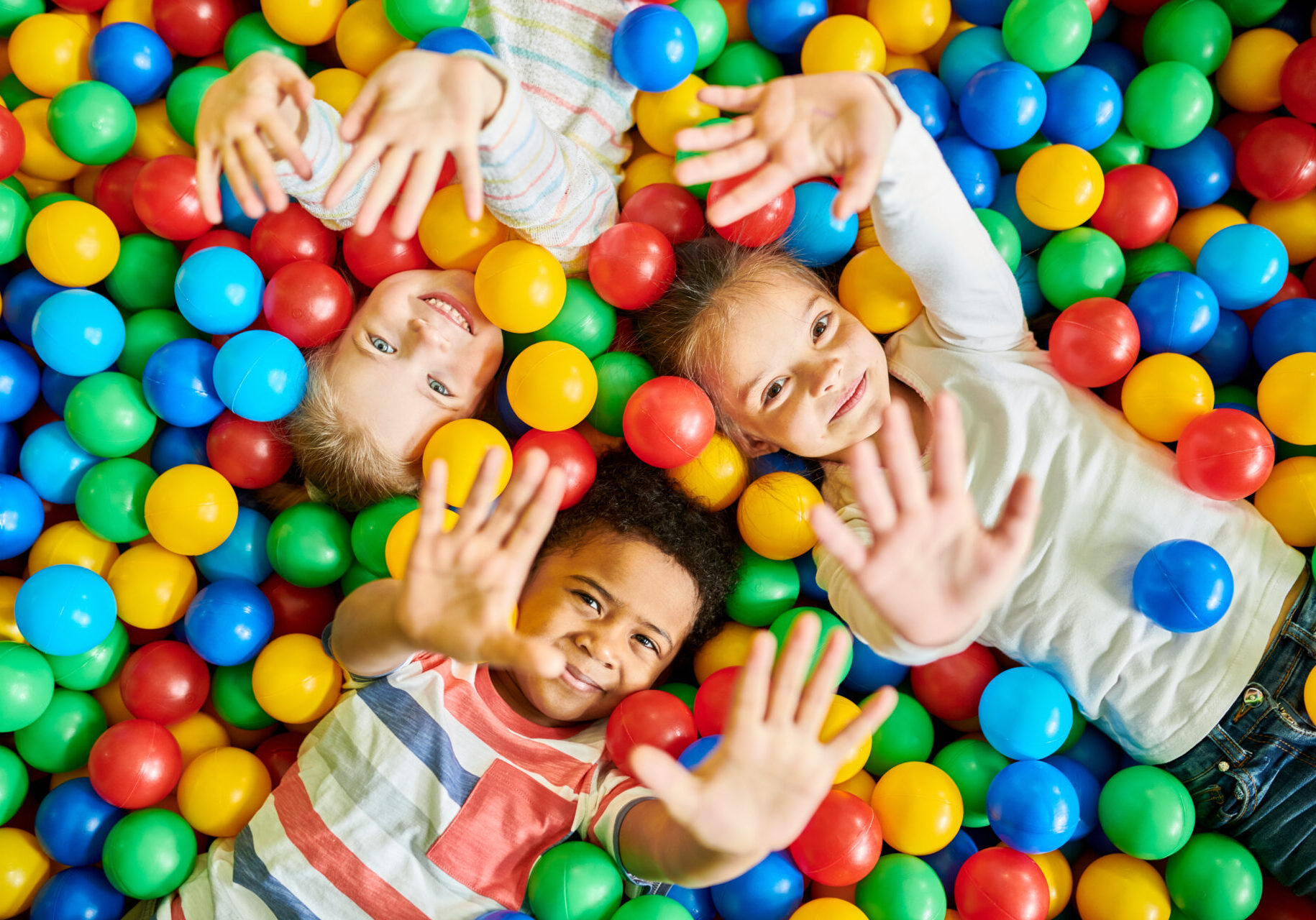 Above view portrait of three happy little kids in ball pit smiling at camera raising hands while having fun in children play center, copy space